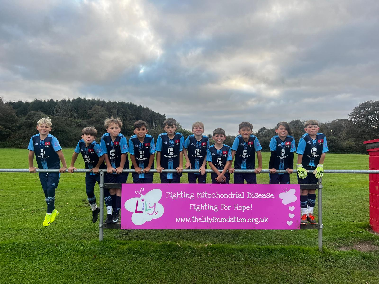 Redruth United youth team lined up behind a Lily Foundation banner reading Fighting Mitochondrial Disease, Fighting For Hope